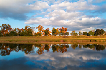 Fototapeta premium Autumn colorful foliage leaves trees reflected in Vltava river with cloudy blue sky. Czech landscape