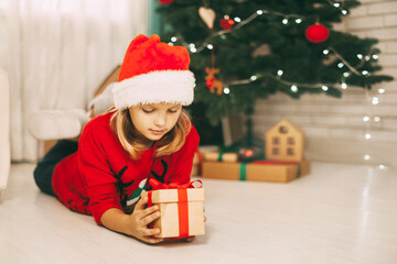 Obraz na płótnie Canvas A blonde girl lies on the floor next to a decorated Christmas tree and holds a gift tied with a red ribbon. The girl unwraps the gift. Christmas and new year concept