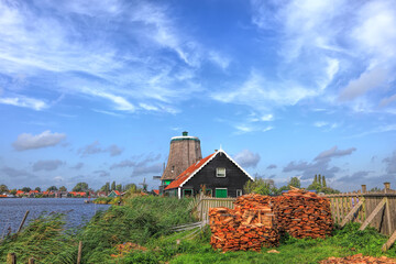 Dutch authentic village on water canal. Old traditional windmills. Stack of wooden boards on a green grass. Gulls fly over water in the blue sky on a sunny day. Netherlands beautiful country landscape