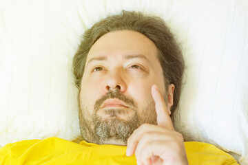 A middle-aged man lies early in the morning on a pillow with his finger raised up. The concept is...
