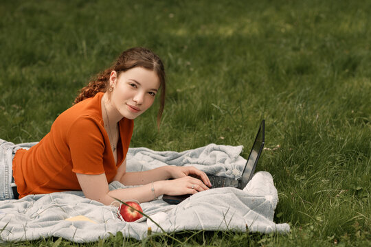 Lifestyle Portait Of Lovely Young Curly Girl College Student. Pretty Woman Looking At The Camera. Female With Laptop Lying On The Green Grass In The Yard. Copy Space.