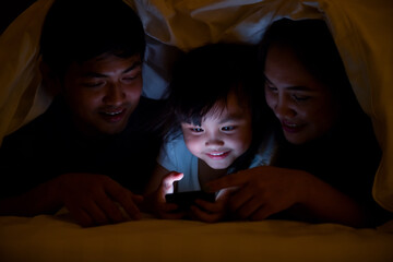 Asian girl children enjoy leisure time with mom and father relax in bedroom using smartphone at night, watch video about teaching content while lying in bed under the blanket together.
