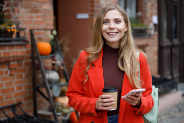 Cheerful young woman wearing coat walking with coffee cup and smartphone. Can use for holiday halloween concept