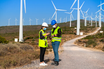 Technical businessman wearing protective face mask working in COVID-19 outbreak after novel coronavirus quarantine and lockdown standing with digital tablet pc on wind power energy station