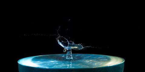 Naklejka na ściany i meble A splash of blue water with a drop flying from above on a black background