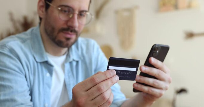 Close up young handsome man holding credit banking card in hands, entering numbers or cvv code in mobile application, shopping in online store, satisfied with easy service usage, quick money transfer.