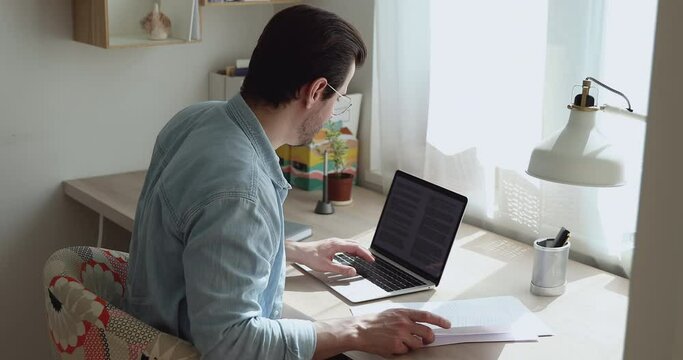 Concentrated 30s young male employee in eyeglasses holding paper documents, checking data making remarks in electronic report on computer, work from home casual freelance quarantine lifestyle workday.