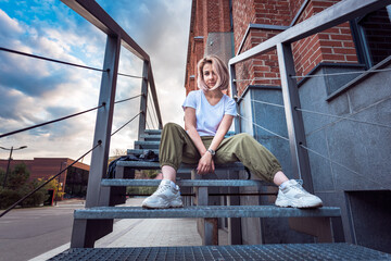 Girl in hip hop clothes sitting on a metal staircase looking at camera with a rebel look in an...