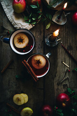 Flat lay with two metal cups with aromatic apple and cranberries tea with cinnamon stick, burning candles, apples and fir branches on old wooden table.