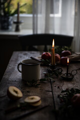 Metal cup with hot apple-cranberry-cinnamon winter tea, lit candles in vintage candleholders , red apples and fir branches on old wooden table in front of window.