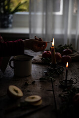 Woman lights a candle on old wooden table with metal cup with hot apple-cranberry-cinnamon winter tea, , red apples and fir branches on it.
