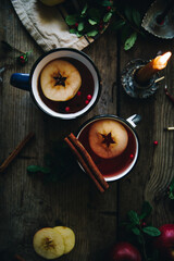 Flat lay with two metal cups with aromatic apple and cranberries tea with cinnamon stick, burning candles, apples and fir branches on old wooden table.