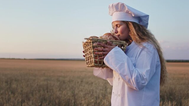 Handsome baker woman smelling the fresh bread from the basket with love in the middle of wheat field she have a pretty smile