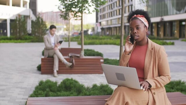  Tracking of young black businesswoman with laptop sitting on bench outside modern business center and talking on mobile phone