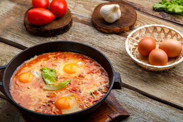 israeli fried eggs shakshuka with cheese in a frying pan near products on a wooden table.