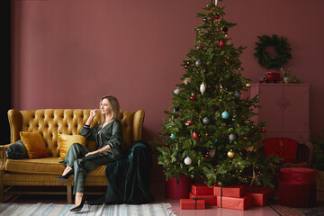 Obraz na płótnie Canvas Model girl in trendy clothes sits on the yellow couch near the Christmas tree in vintage interior