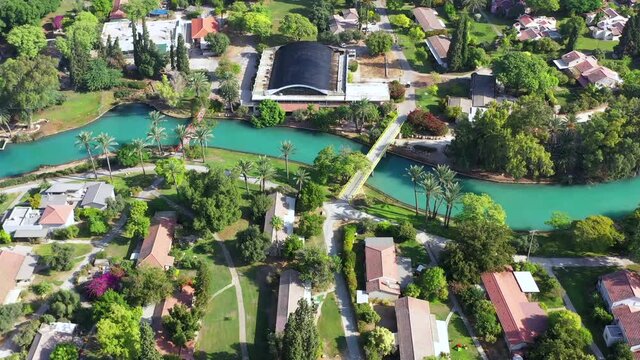 Aerial pass over Kibbutz Nir David with Asi river channel turquoise water dividing east and west side riverside houses and palm trees, Israel.