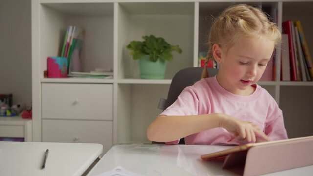 Cute elementary school girl using digital tablet for learning and thinking with her hand on cheek sitting at desk at home, tracking medium shot