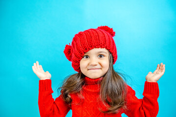 Portrait of a little curly-haired girl in a knitted red hat in winter. little girl with dark hair in a red knitted hat and sweater smiling on a blue background isolate, place for text. Winter clothes