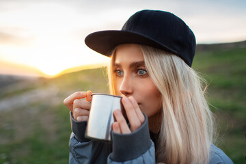 Close-up portrait of young blonde girl with black cap drinking hot tea from steel mug in background...