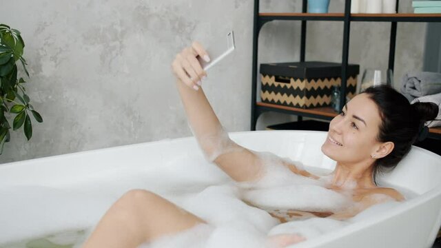 Joyful young woman is posing for camera with funny faces taking selfie in bathtub having fun at home. Modern devices and photography concept.