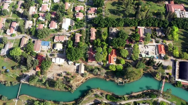 Aerial pass over Kibbutz Nir David with Asi river channel turquoise water dividing east and west side riverside houses and palm trees, Israel.