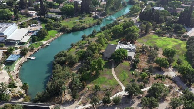 Aerial pass over Kibbutz Nir David with Asi river channel turquoise water dividing east and west side riverside houses and palm trees, Israel.