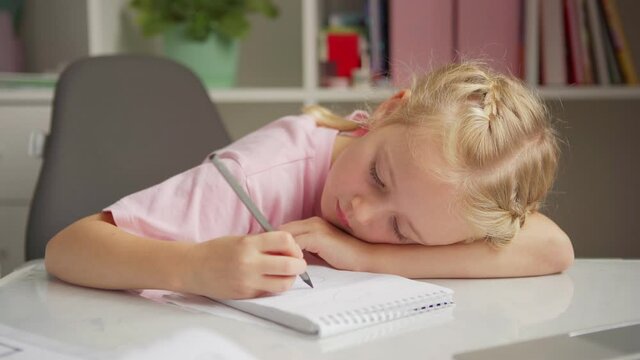 Tired elementary school girl lying down on desk and drawing hearts in her notepad while doing homework at home