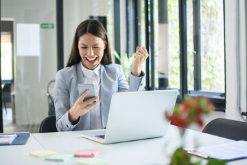 Happy excited woman holding phone and triumphing with fist up in office.