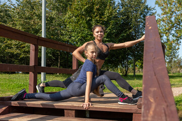 Mother and daughter stretching legs together on top of wooden staircase in public park on sunny summer morning. Family outdoor sports theme.