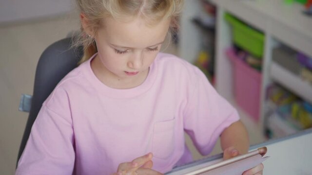 From above tilt up shot of cute elementary school girl using digital tablet for studying and looking up dreamily while doing homework sitting at desk at home