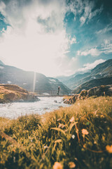 A young woman standing on a bridge in Grossglockner national park surrounded by mountains and alpine stream on a sunny day - wide shot