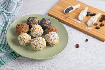 Homemade energy balls with dried fruits, nuts, sesame seeds, cocoa, almonds and poppy seeds on a plate, spices in wooden spoons, light background. Vegan and vegetarian raw appetizer or food. Close up