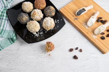 Home-made energy balls with dried fruits, nuts, sesame seeds, cocoa, almonds and poppy seeds on a black plate, spices in wooden spoons. Vegan raw appetizer or food. Close up.