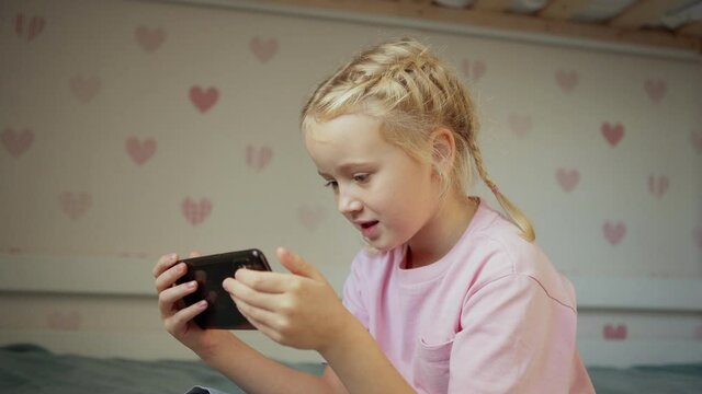 Tilt up side view medium shot of beautiful blonde little girl watching cartoons or online lesson on cell phone sitting on bed at home
