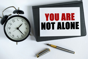 On the table there is a clock, a pen, a notebook and a card on which the text is written - YOU ARE NOT ALONE