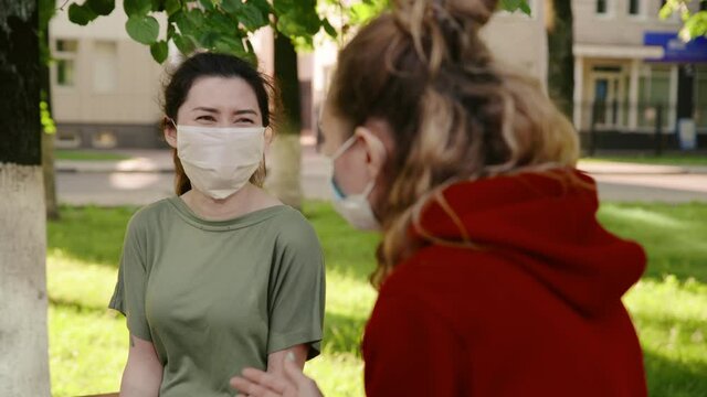 Young pretty girls talk to each other in protective masks from the coronovirus and laughting. Woman tells her friend a funny story. New life after covid-19 epidemic. Concept of pandemic, corona.