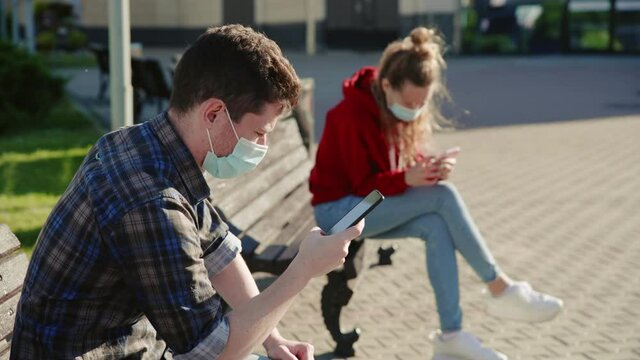 Girl And Guy Socially Distance Themselves From Each Other. People Communicate Through Their Smartphone Or Mobile Phone And Sitting On Different Benches Outdoor In Medical Masks. Keep Safe Distance.