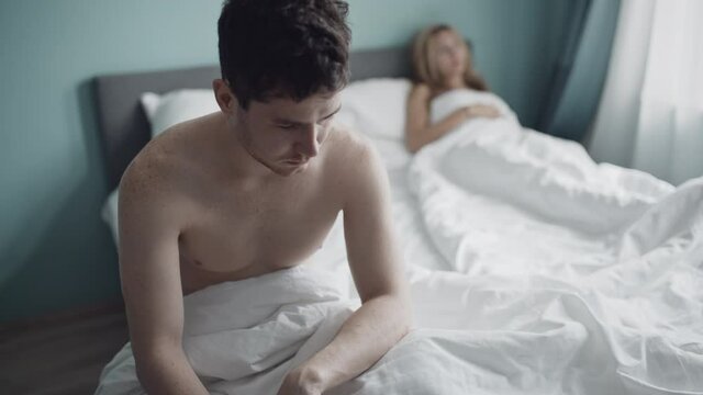 Nervous guy upset sitting on edge of bed because of problems with sex. Young caucasian couple having relationship anxiety. Family conflict. Relationship crisis in couple. Erectile disfunction concept.