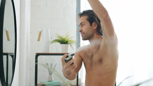 Slow motion of good-looking young man applying deodorant on armpits skin looking at mirror in bathroom during morning routine. People and lifestyle concept.