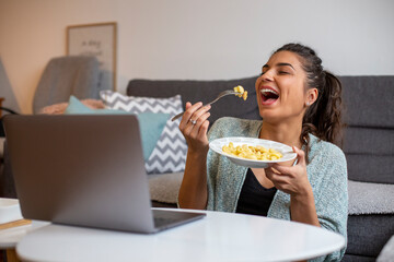 A smiling young woman is eating pasta and working on a laptop in the living room