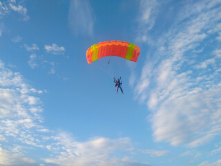 A skydiver with a bright orange sports parachute wing flies before landing in the summer against a blue sky background close up