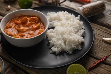 Delicious chicken curry dish with rice on black round plate served with lime on rustic wooden background. Tasty asian dinner concept. Hot spicy dish.