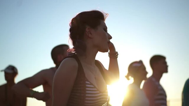 Pretty asian woman dancing at sunset light among the crowd of people at rock concert festival. Concept of youth culture and entertainment. Music fans enjoying summer gig.