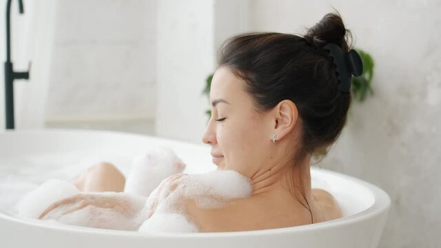 Pretty carefree girl is enjoying warm relaxing bath touching gentle white foam in bathroom. Beauty activities and modern lifestyle concept.