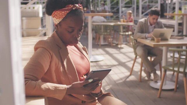 Tracking shot of serious African-American businesswoman with tablet sitting in outdoor patio of cafe and typing on tablet