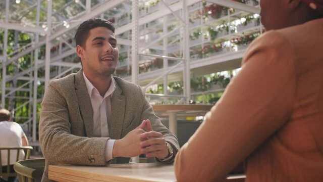  Low angle shot of happy businessman sitting at table in outdoor cafe patio and having meeting with black businesswoman. He is talking and shaking her hand