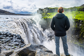 Young man back looking at Dettifoss waterfall on rocks water flowing mist spraying cloudy day in...