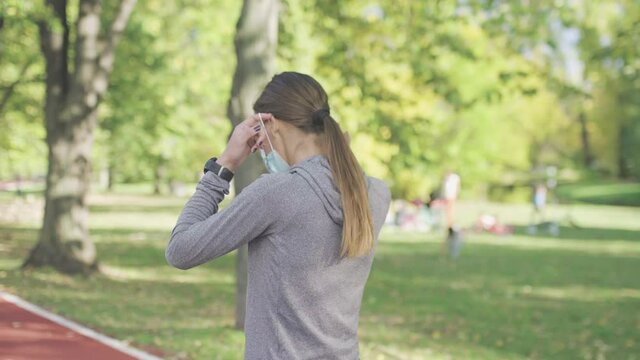 Runner girl walking on an all weather synthetic track surface in the park. Wearing protective face mask, and taking it off. Sport during pandemic