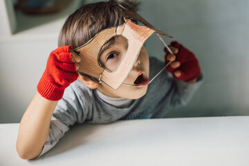 Cute kid in disguise with DIY  cardboard superhero mask at home. Boy with creative Halloween...